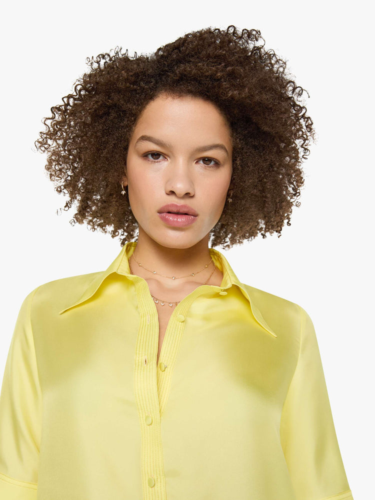 Close up view of a woman in a light yellow shirt with collared neckline with an extra-long button placket, elbow-length short sleeves and a loose, boxy fit.