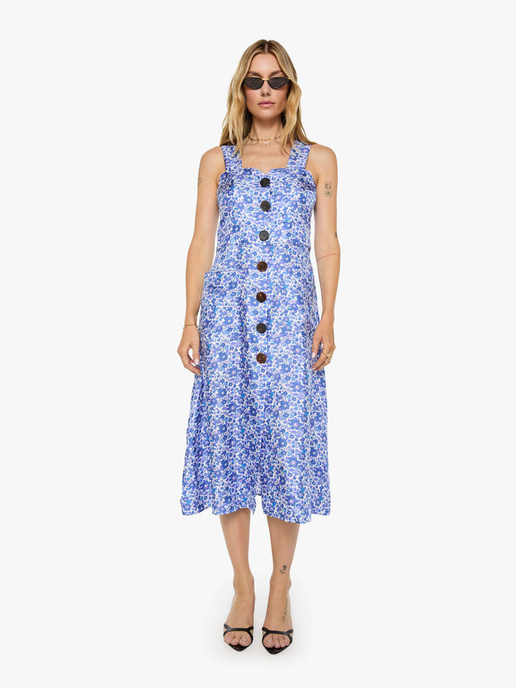 Front view of a woman in a blue and white floral print dress with wide straps, a scoop neck, black buttons down the front and a calf-length hem.