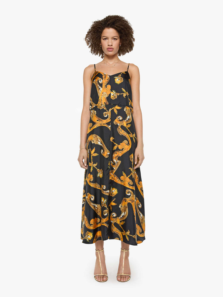 Front view of a woman in a black and gold prints with classic silhouettes, featuring a subtle V-neck, thin spaghetti straps, semi-fitted waist and a flowy ankle-length skirt.