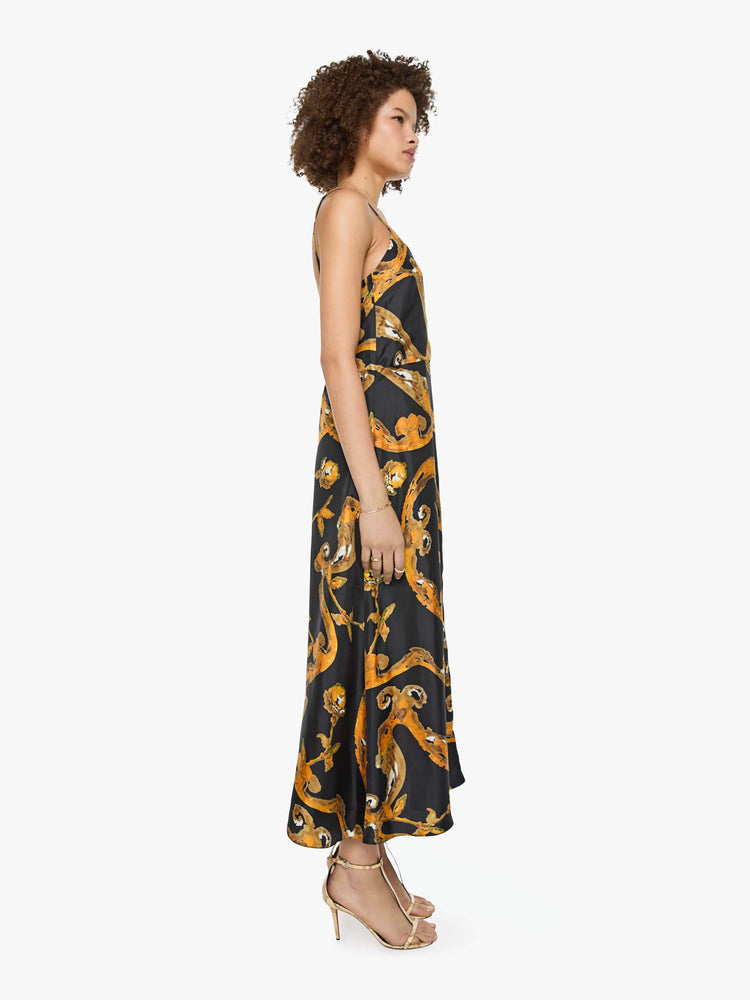 Side  view of a woman in a black and gold prints with classic silhouettes, featuring a subtle V-neck, thin spaghetti straps, semi-fitted waist and a flowy ankle-length skirt.