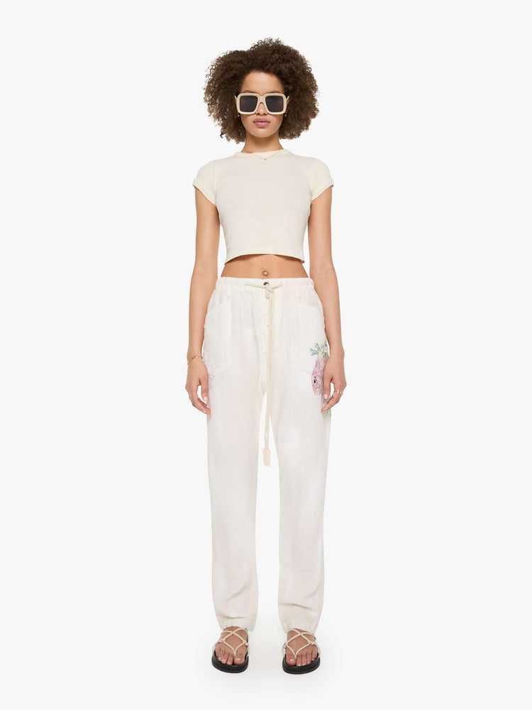 Front view of a woman in white with a hand-drawn koi fish screen-printed on the left side pocket, the pants have oversized patch pockets, an extra-long inseam that puddles at the hem and a woven belt that ties.