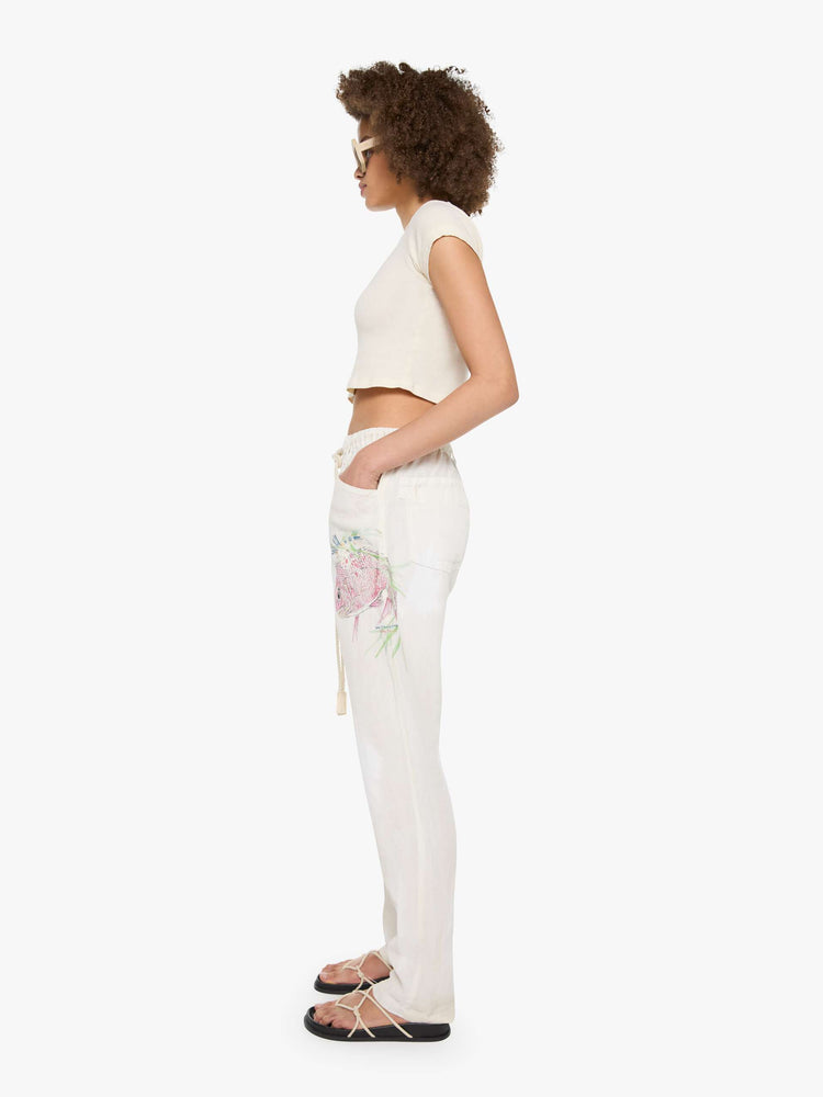 Side view of a woman in white with a hand-drawn koi fish screen-printed on the left side pocket, the pants have oversized patch pockets, an extra-long inseam that puddles at the hem and a woven belt that ties.