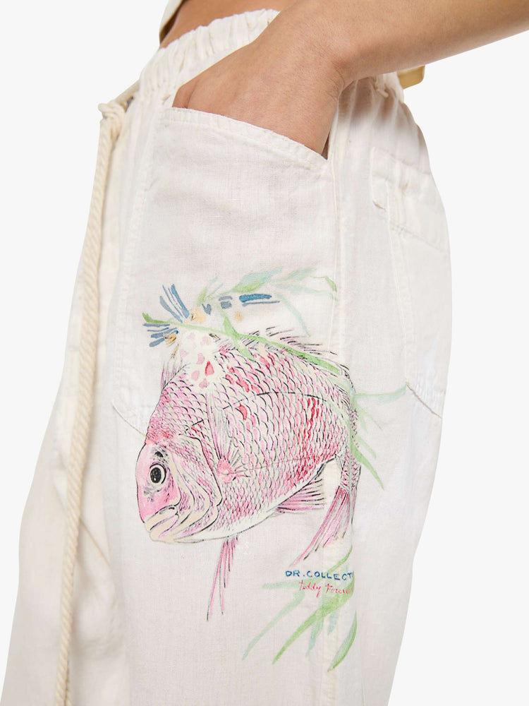 Close up view of a woman in white with a hand-drawn koi fish screen-printed on the left side pocket, the pants have oversized patch pockets, an extra-long inseam that puddles at the hem and a woven belt that ties.
