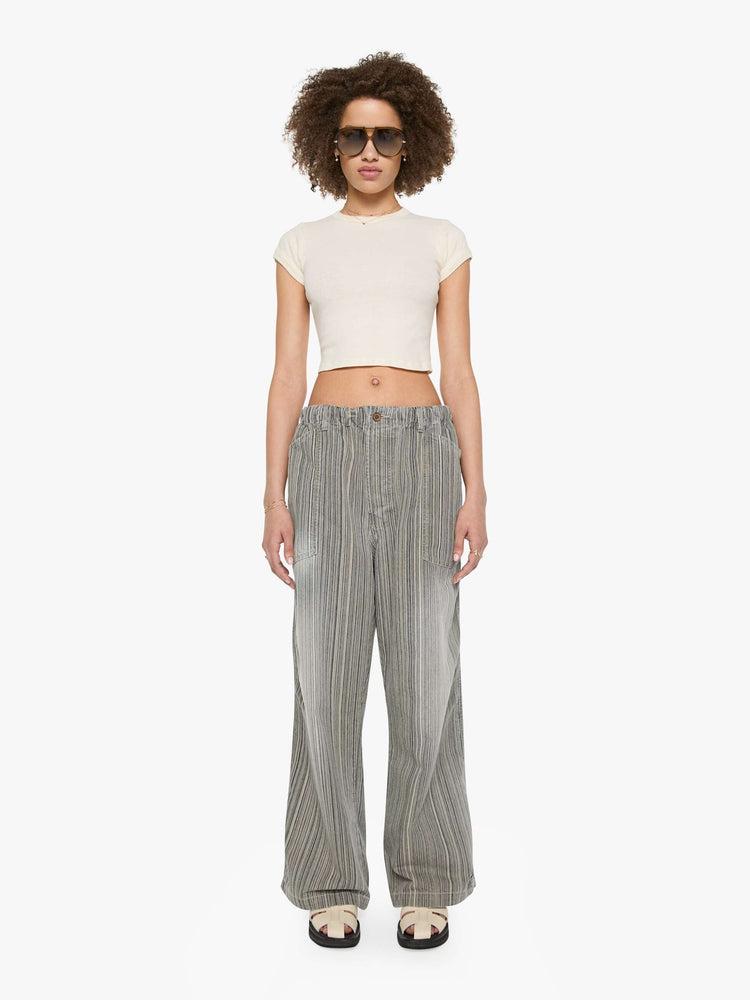 Front view of a woman in faded grey with vertical stripes pants with a mid rise with oversized pockets, an extra-long inseam that puddles at the hem and a colorful woven belt that ties.