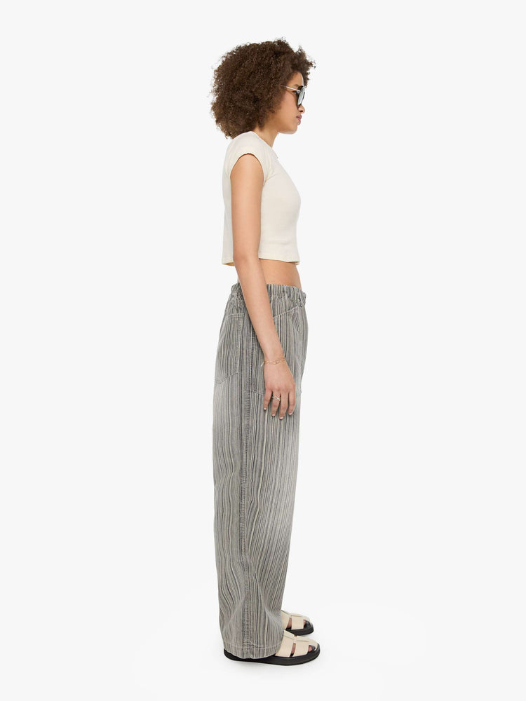 Side view of a woman in faded grey with vertical stripes pants with a mid rise with oversized pockets, an extra-long inseam that puddles at the hem and a colorful woven belt that ties.