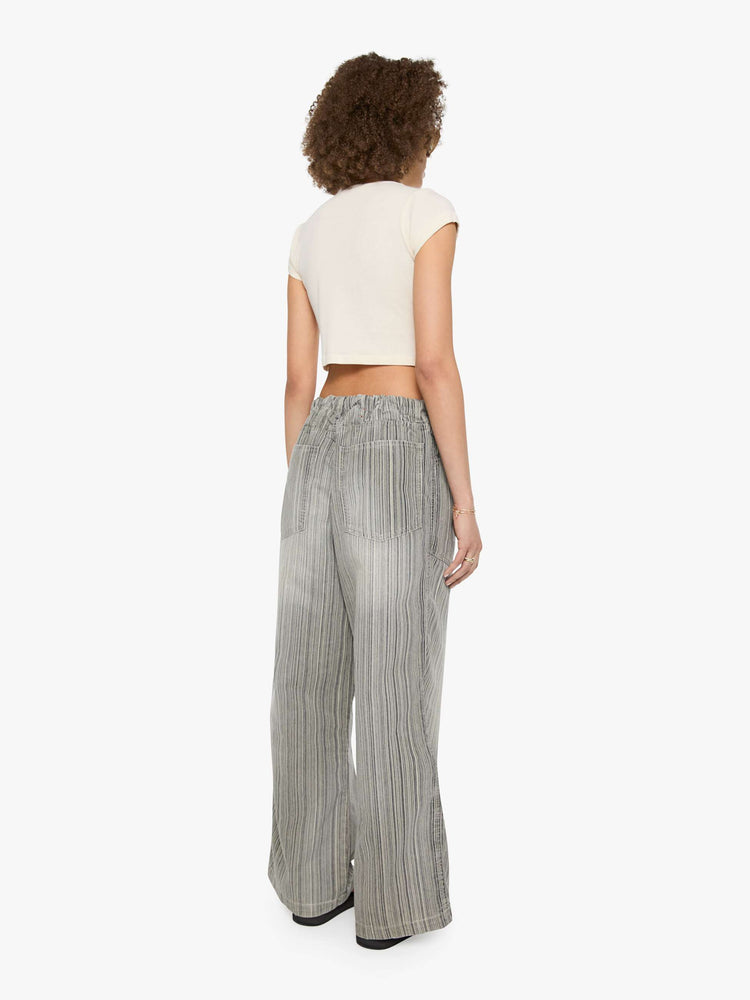 Back view of a woman in faded grey with vertical stripes pants with a mid rise with oversized pockets, an extra-long inseam that puddles at the hem and a colorful woven belt that ties.