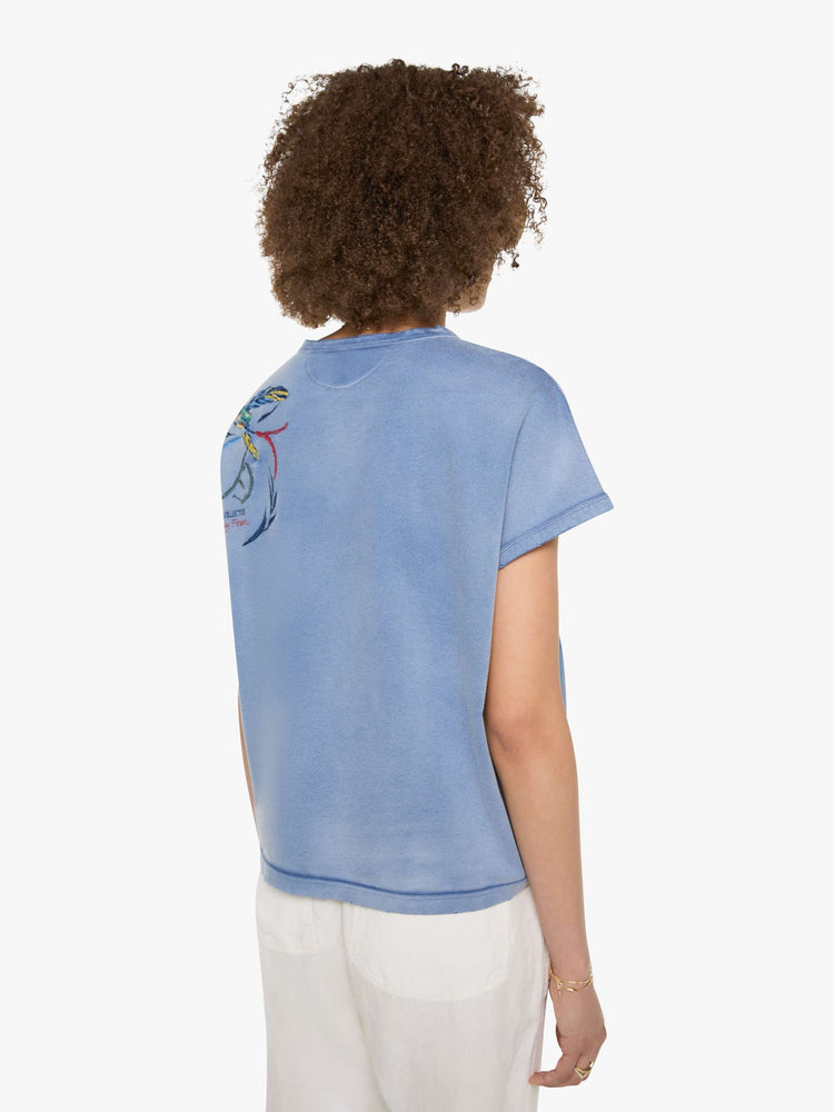 Back view of a woman in a sun-faded blue  tee with a colorful chain-stitched hummingbird and text on the back left shoulder.