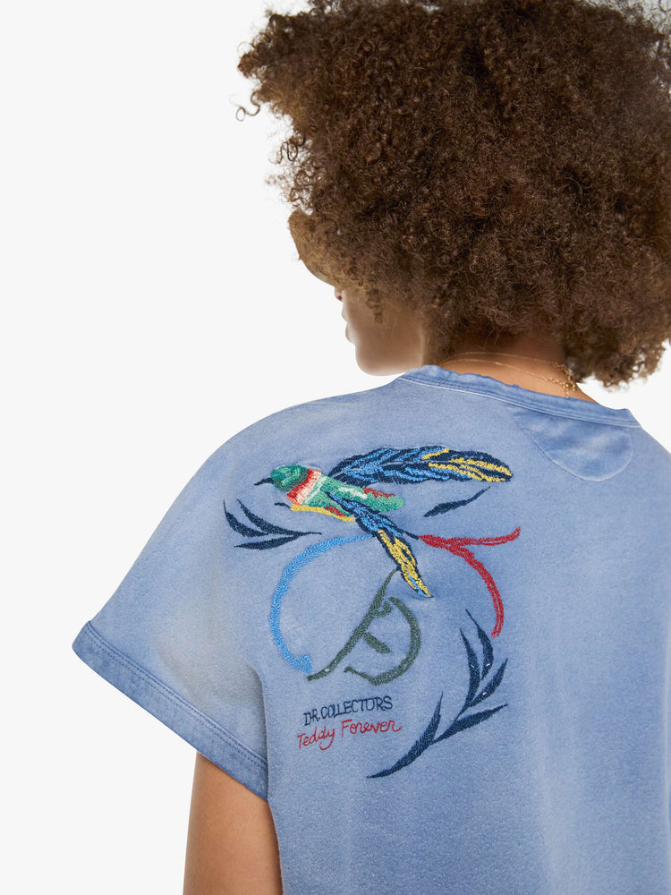 Back close up view of a woman in a sun-faded blue  tee with a colorful chain-stitched hummingbird and text on the back left shoulder.