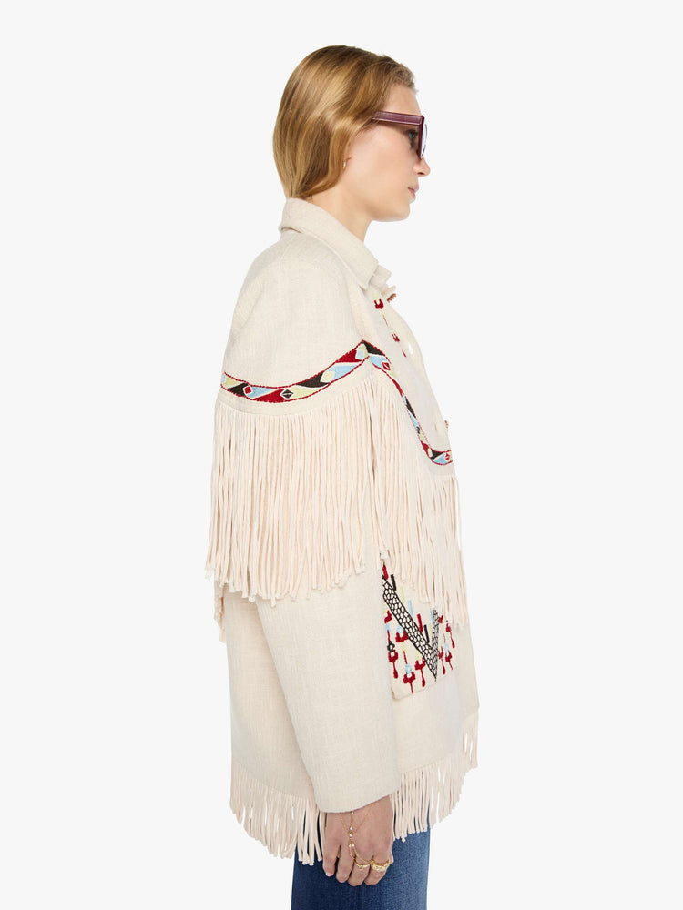 Side view of a woman in a cream jacket with decorative embroidery, patch pockets, fringe at the chest and hem and a boxy fit.