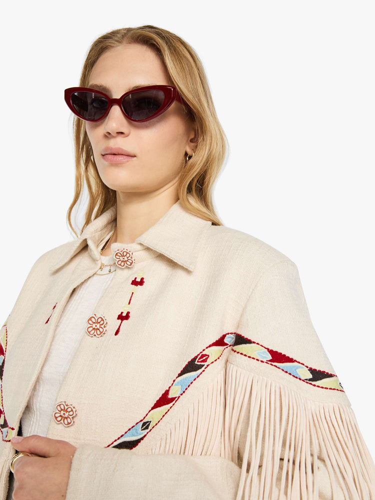 Close up view of a woman in a cream jacket with decorative embroidery, patch pockets, fringe at the chest and hem and a boxy fit.
