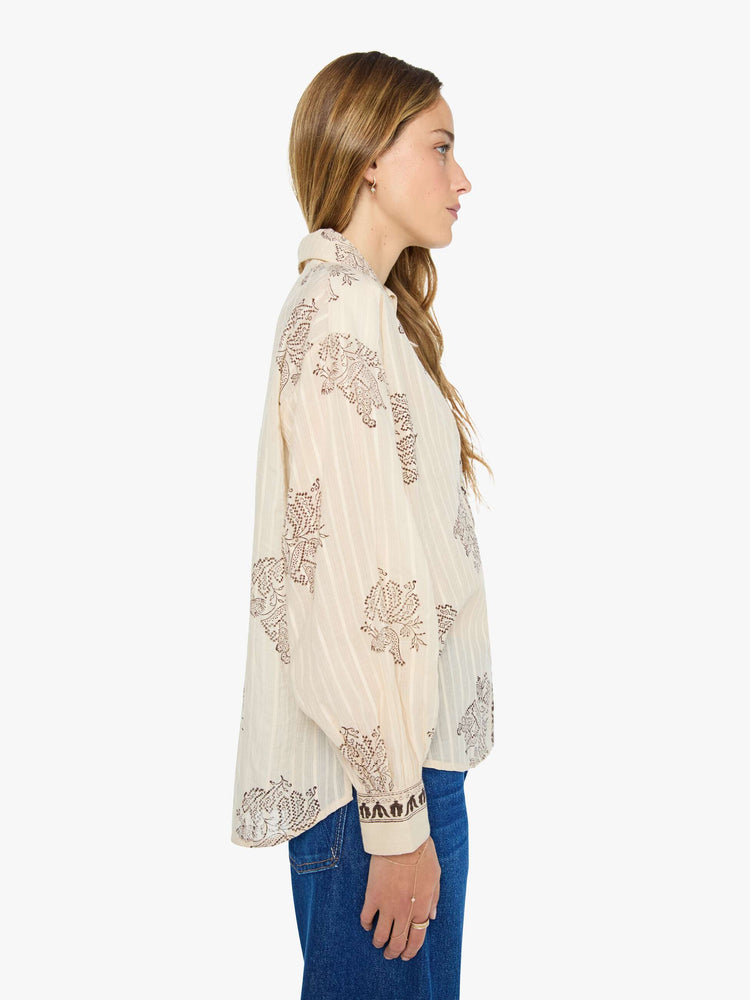 Side view of a woman in cream button up with an oversized floral print, and features a stacked collar, long balloon sleeves and ruffles throughout for a loose, flowy fit.