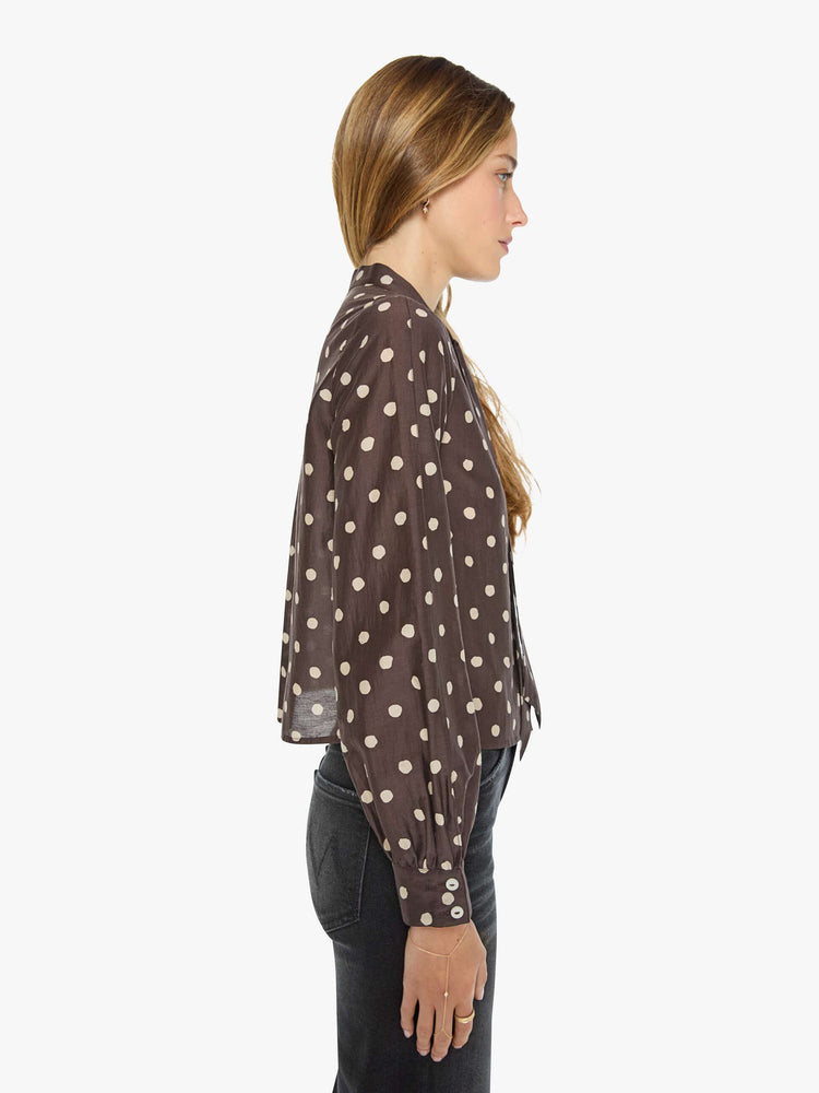 Side view of a woman in a mahogany with cream polka dots shirt with a collared neck that ties, long balloon sleeves and a cropped fit.