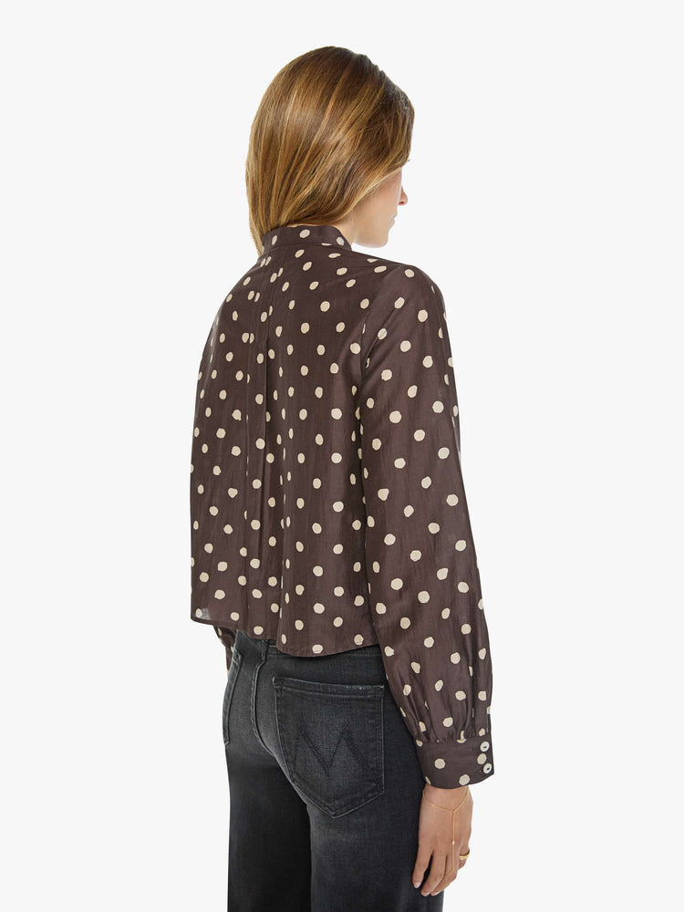 Back view of a woman in a mahogany with cream polka dots shirt with a collared neck that ties, long balloon sleeves and a cropped fit.