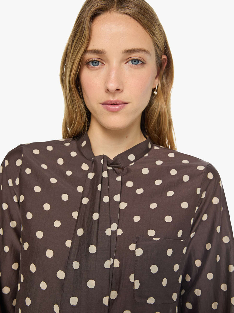 Close up view of a woman in a mahogany with cream polka dots shirt with a collared neck that ties, long balloon sleeves and a cropped fit.
