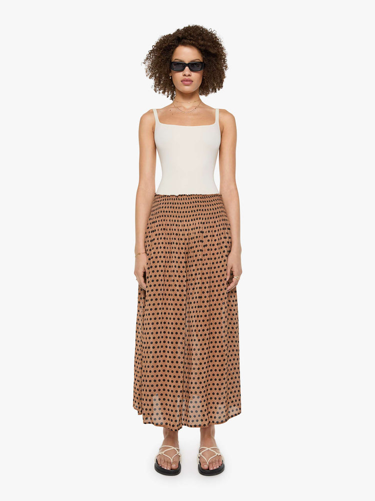 Front view of a woman in a tan with black polka dots maxi skirt has a smocked waistband and a loose, flowy fit.