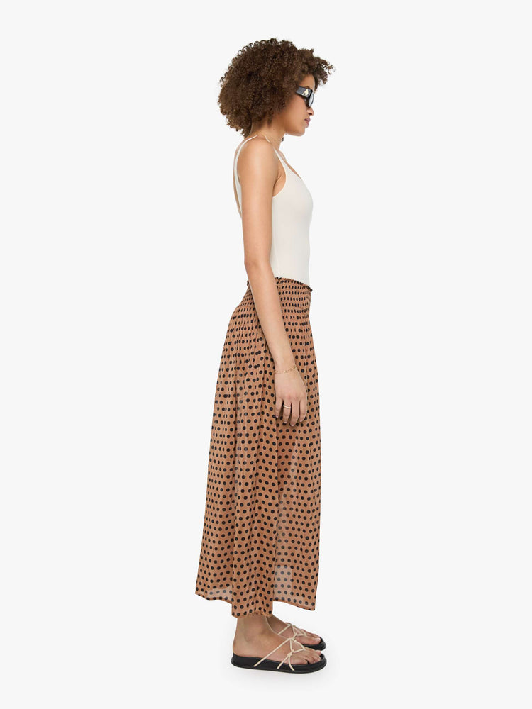 Side view of a woman in a tan with black polka dots maxi skirt has a smocked waistband and a loose, flowy fit.