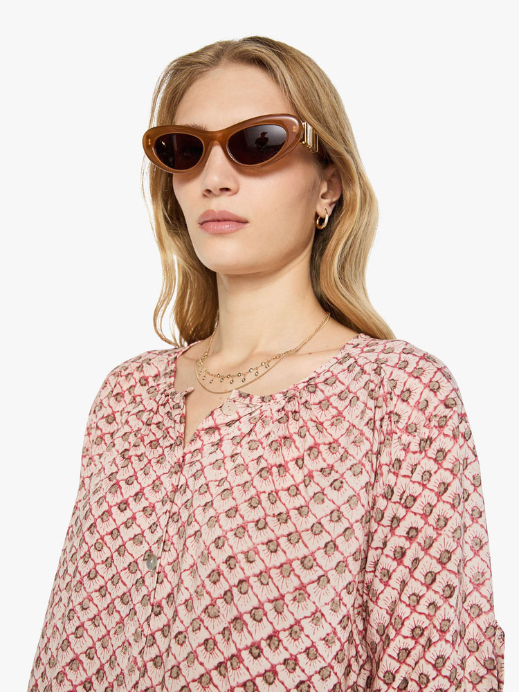 Close up view of a woman in a light pink with a delicate maroon print top features a deep V-neck, buttons down the front, short sleeves and is slightly pleated under the collar for a loose, flowy fit.