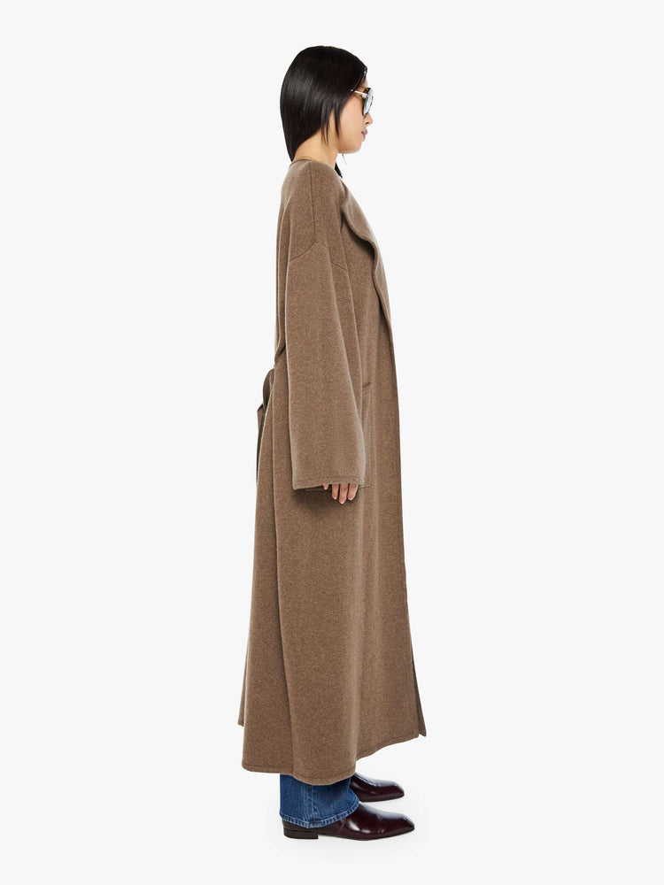 Side view of a woman in a chocolate brown trench coat with an oversized collar, drop shoulders, a tied waist and calf-length hem.