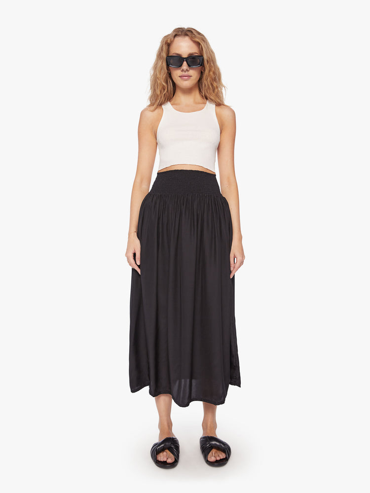 Front view of a woman wearing a black skirt featuring a wide elastic waistband and a flowy fit, paired with a cropped white tank top.