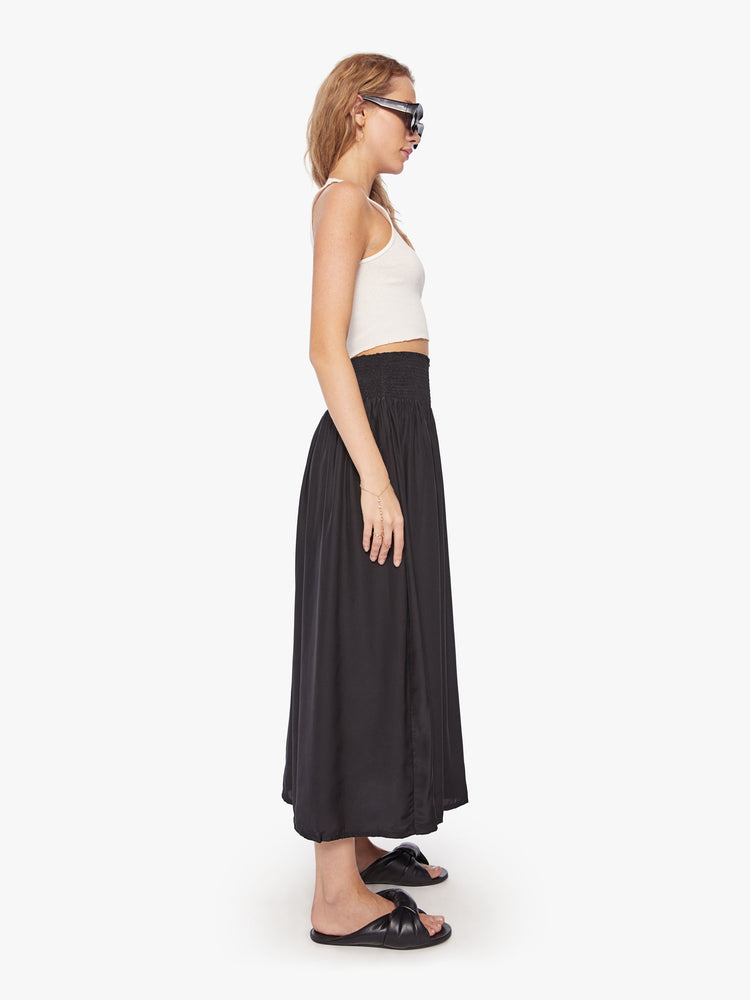 Side view of a woman wearing a black skirt featuring a wide elastic waistband and a flowy fit, paired with a cropped white tank top.