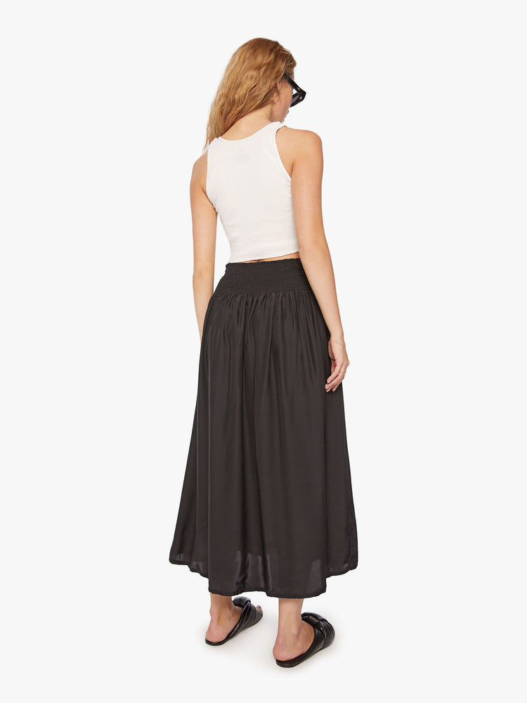 Back view of a woman wearing a black skirt featuring a wide elastic waistband and a flowy fit, paired with a cropped white tank top.