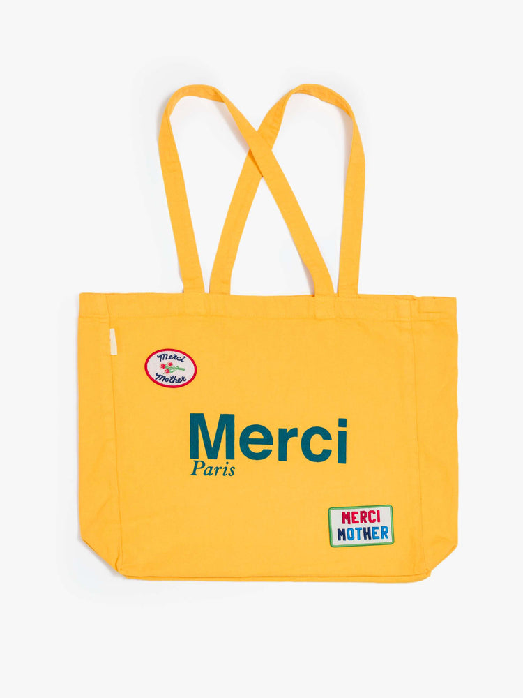 Flat view a bright yellow tote with bold flocked text in blue, finished in a raised, velvety texture, and colorful patches at the bottom right and top left corners.