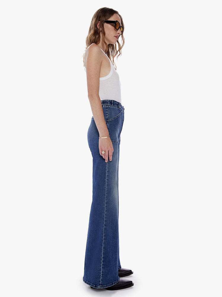 Side full body view of a woman in an inspired wide leg pant with a high rise, patch pockets, long 32 inch inseam, and loose fit made from denim with a touch of stretch in a dark blue wash with subtle fading and whiskering