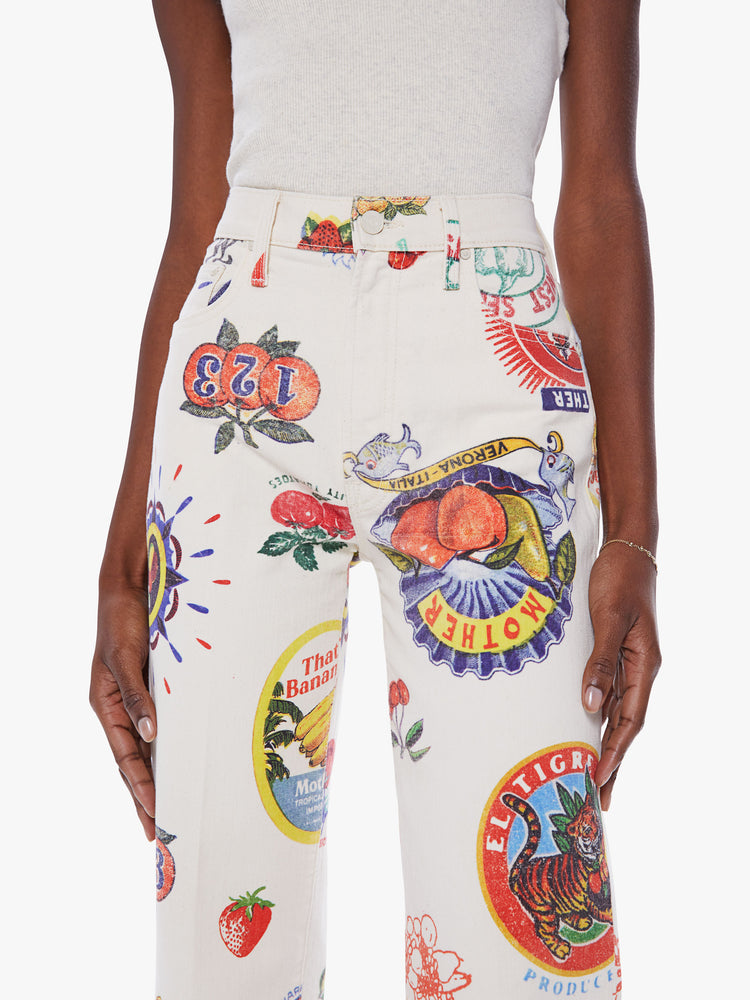 Close up view of a woman's high-waisted jeans with a wide straight leg, zip fly and clean ankle-length inseam in a white hue with faded fruit sticker graphics and doodles.