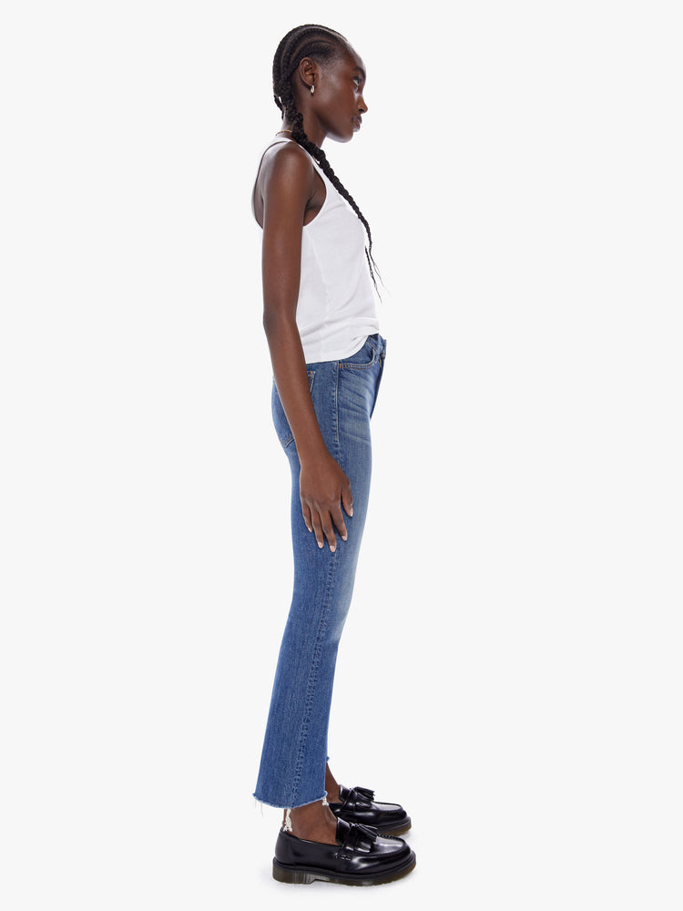 Side full body view of a woman in a high rise flare frayed hem and an inseam that hits just below the ankle is a mid blue wash with fading and whiskering