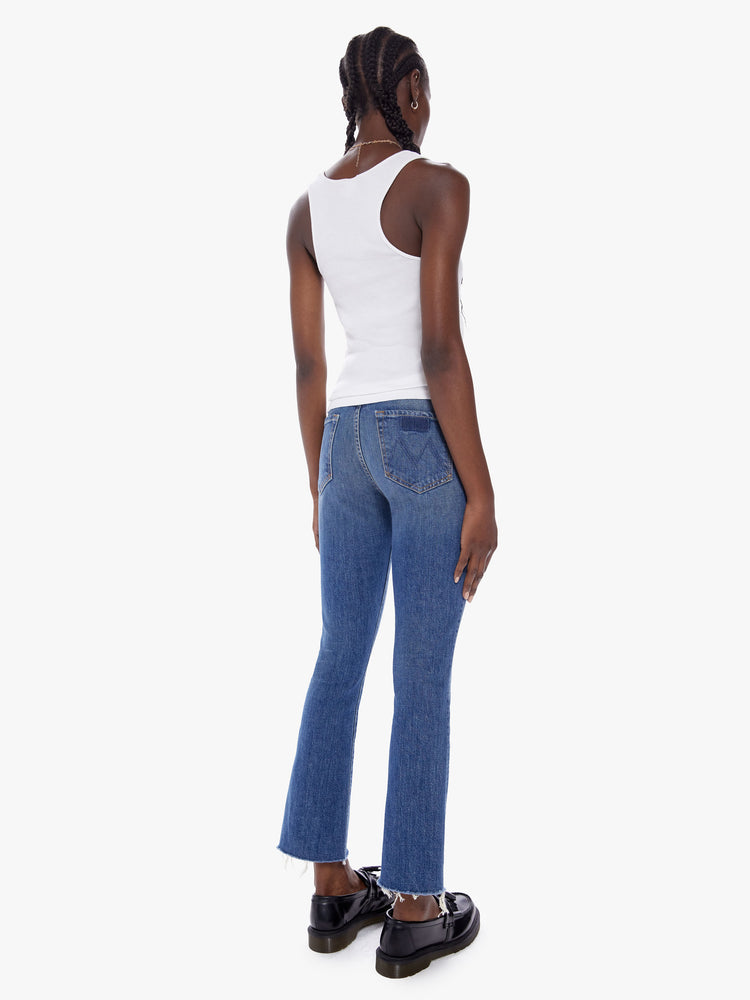 Back full body view of a woman in a high rise flare frayed hem and an inseam that hits just below the ankle is a mid blue wash with fading and whiskering