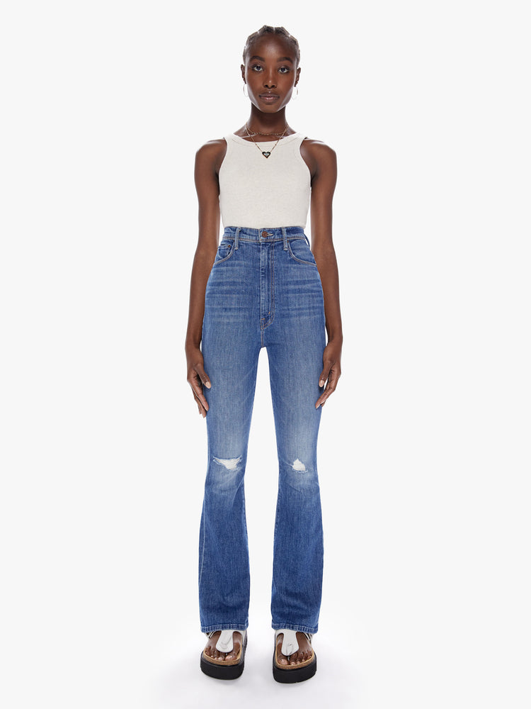 Front full body view of a woman in an ultra high waisted jean with a flared leg, zip fly and a long 32 inch inseam cut from a touch of stretch in a mid blue wash with whiskering fading and distressed details