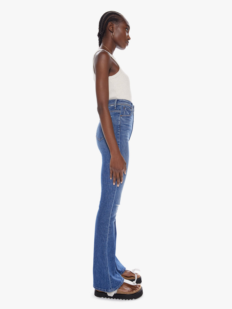 Side full body view of a woman in an ultra high waisted jean with a flared leg, zip fly and a long 32 inch inseam cut from a touch of stretch in a mid blue wash with whiskering fading and distressed details
