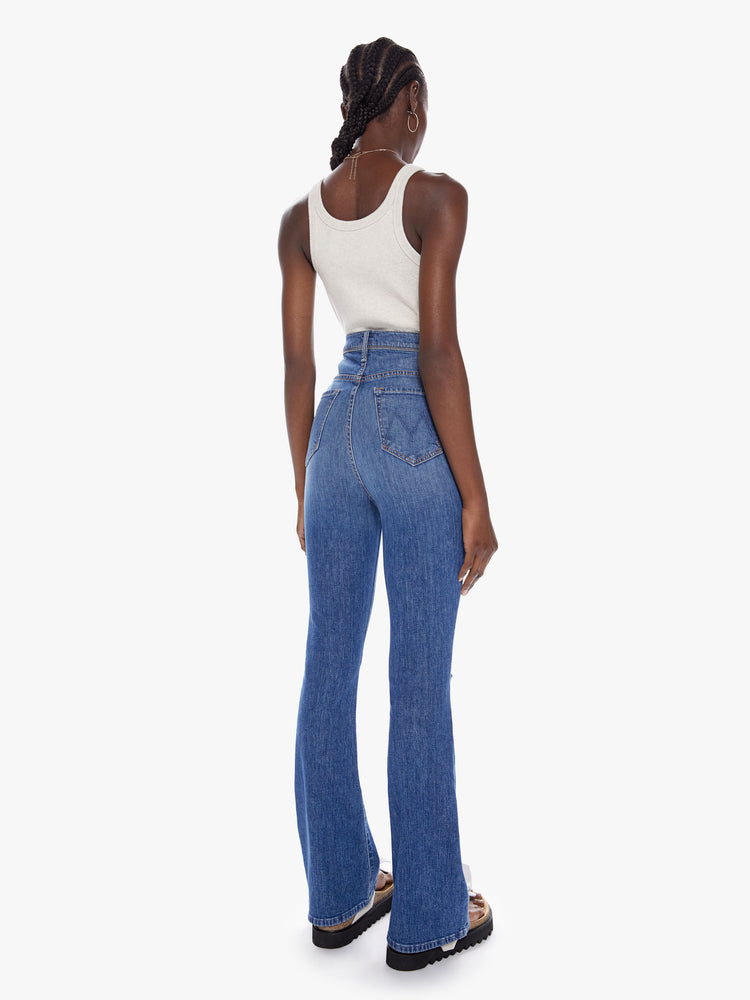 Back full body view of a woman in an ultra high waisted jean with a flared leg, zip fly and a long 32 inch inseam cut from a touch of stretch in a mid blue wash with whiskering fading and distressed details