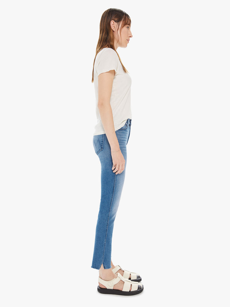 Side view of a womens medium blue wash jean featuring a mid rise, slim straight leg, and a raw cut split hem.