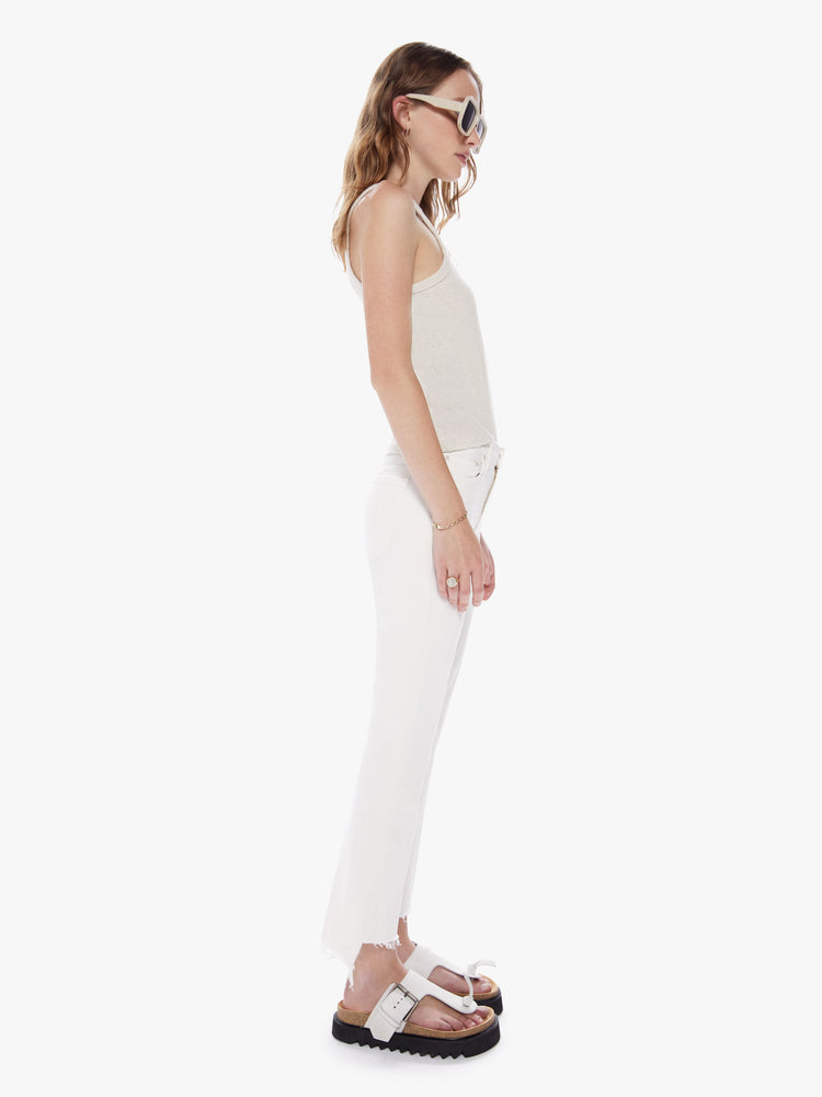 Side full body view of a woman in a high waisted bootcut hits at the ankle with a frayed step-hem cut from comfortable stretch fabric in an off white hue with tonal hardware