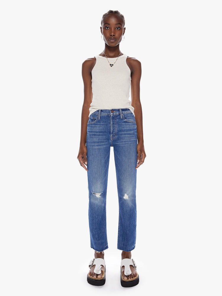 Front full body view of a woman in a high rise jean with a straight leg, button fly, ankle length inseam and a frayed hem made with a touch of stretch in a mid blue wash with whiskering, fading and distressed details