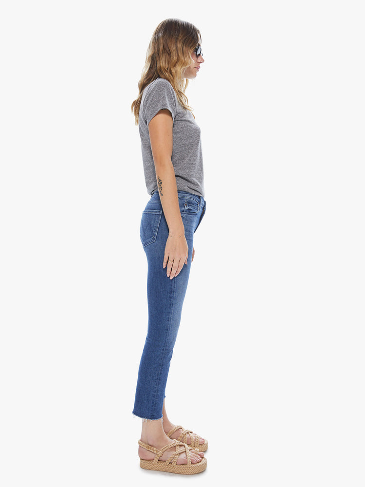 Side view of a woman wearing medium blue wash jeans featuring a mid rise and a frayed cropped hem.