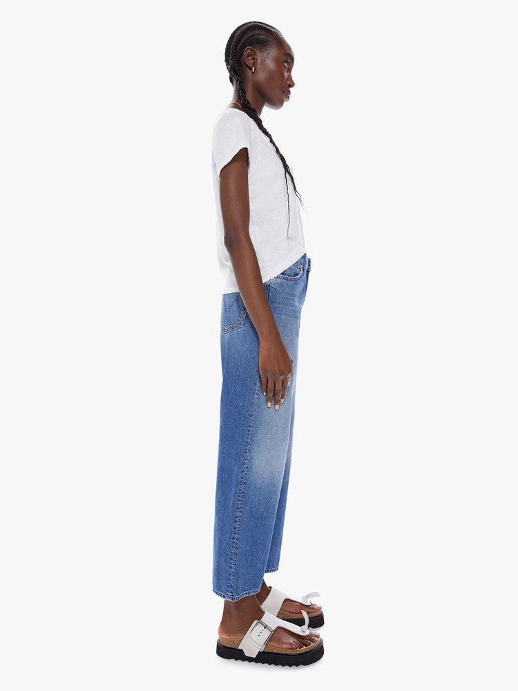 Side full body view of woman in a high waisted jean with loose wide leg and an ankle length inseam cut from a recycled cotton blend denim in a midblue wash with whiskering and fading at the knees