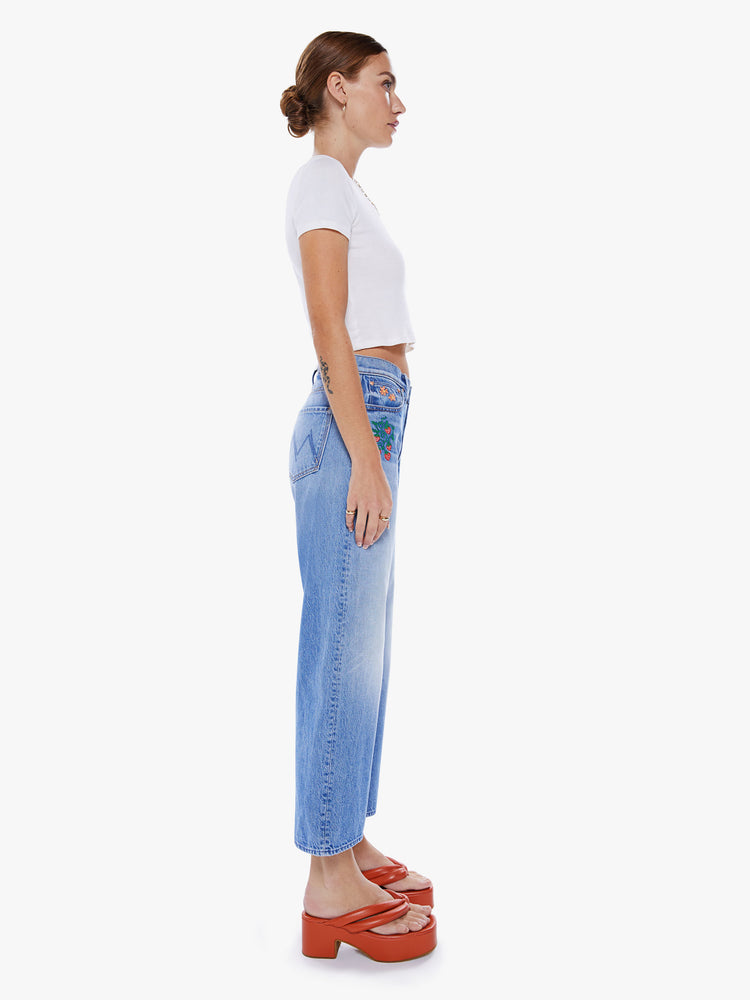 Side view of a woman high-waisted jeans with a loose wide leg and an ankle-length inseam in a mid blue wash and colorful garden-inspired embroidery throughout.