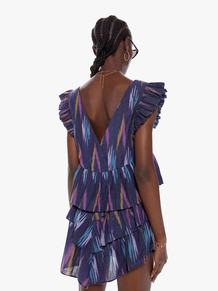 Back view of a woman in a square neck blouse with short ruffled sleeves and a low back made from 100% cotton in a dark blue with a brush stroke inspired pattern in lavender, mustard, and baby blue