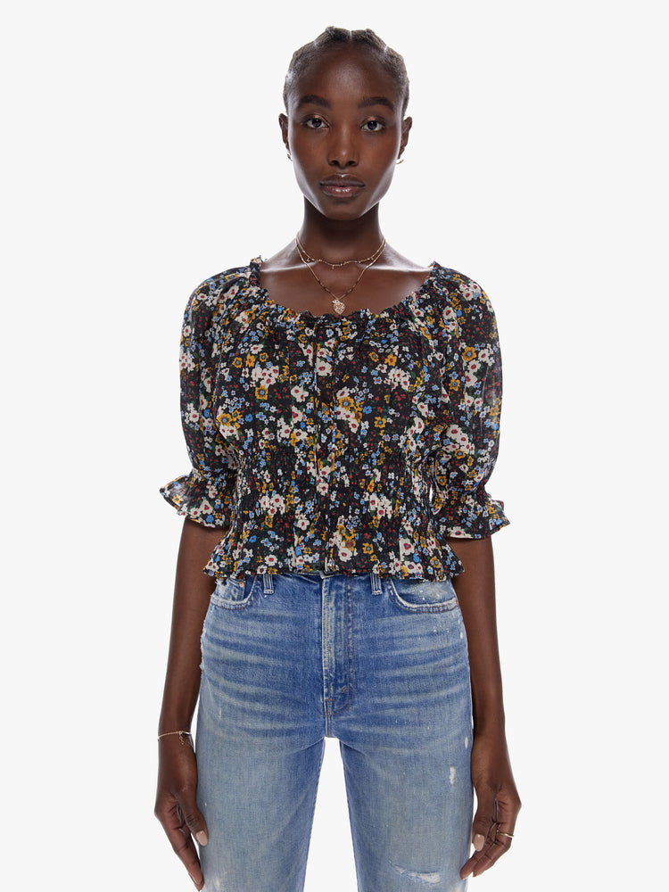 Close front view of a woman in a cropped blouse with a gathered scoop neck that ties, 3/4 - length balloon sleeves, a smocked elastic waist and ruffled hems made from 100% cotton in a contrast floral print in black, blue, yellow, red and white
