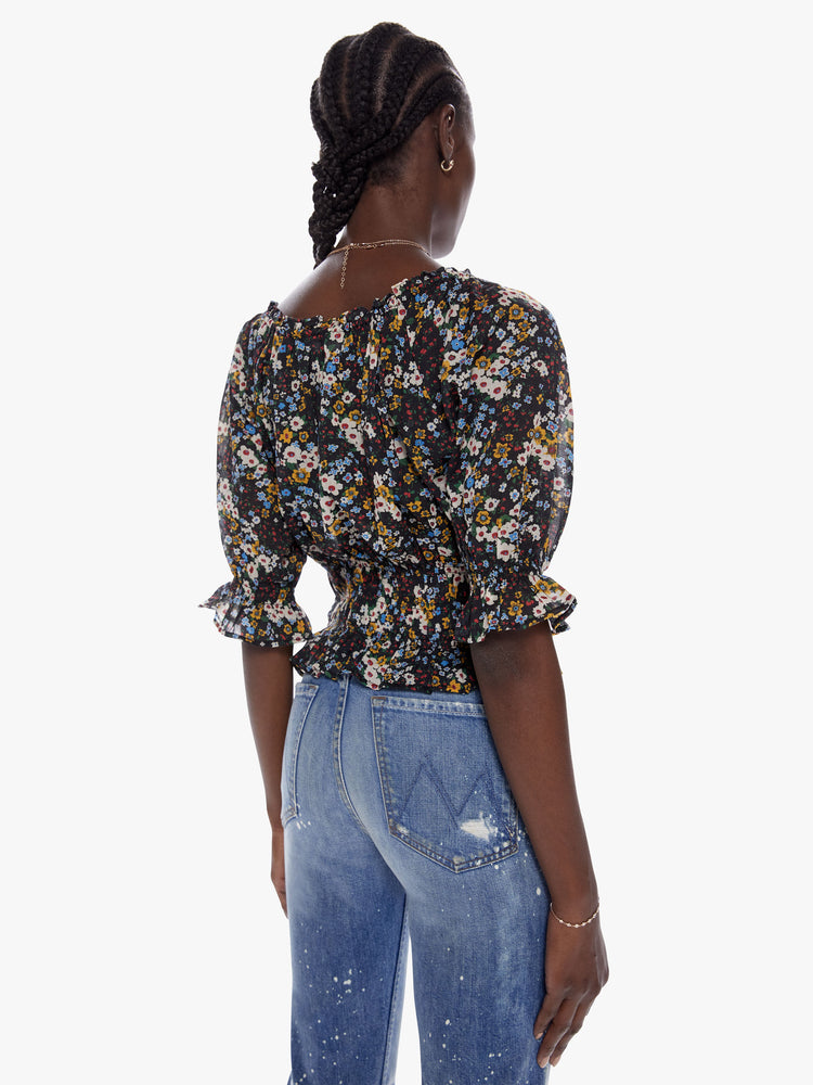 Back view of a woman in a cropped blouse with a gathered scoop neck that ties, 3/4 - length balloon sleeves, a smocked elastic waist and ruffled hems made from 100% cotton in a contrast floral print in black, blue, yellow, red and white