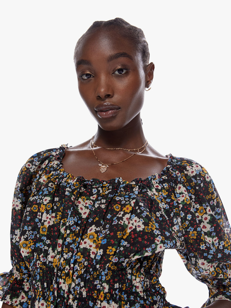 Close up view of a woman in a cropped blouse with a gathered scoop neck that ties, 3/4 - length balloon sleeves, a smocked elastic waist and ruffled hems made from 100% cotton in a contrast floral print in black, blue, yellow, red and white