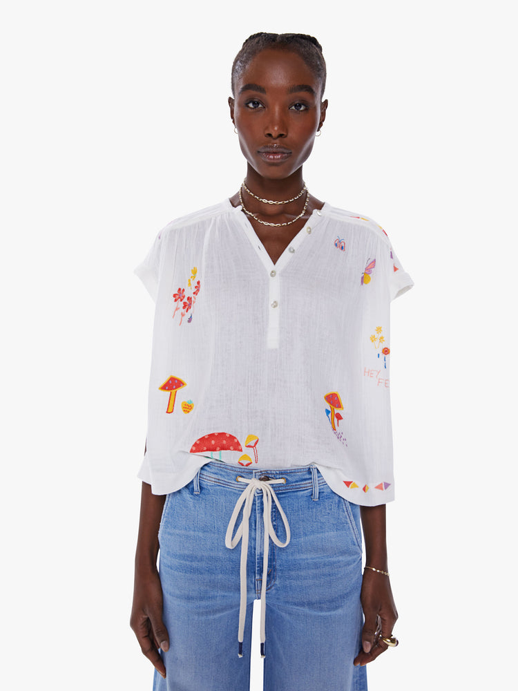 Front view of woman's blouse with a buttoned V-neck, rolled short sleeves, gathered seams at the shoulders and a boxy fit in semi-sheer white with embroidered flowers, mushrooms and butterflies.