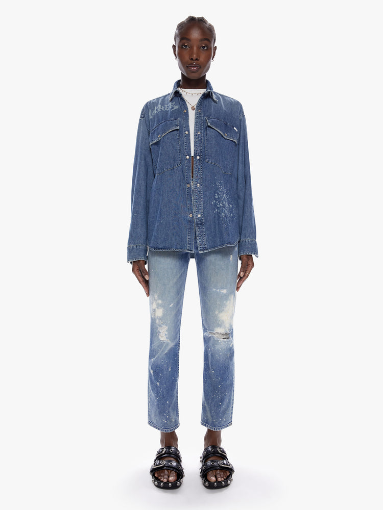 Full body view of woman in an oversized denim button up with long sleeves, oversized patch pockets, a curved hem and snaps down the front made from recycled cotton blend in a midblue wash with mothers name beached in death metal inspired front on the chest and back