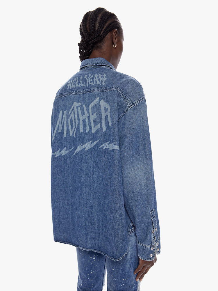 Back view of woman in an oversized denim button up with long sleeves, oversized patch pockets, a curved hem and snaps down the front made from recycled cotton blend in a midblue wash with mothers name beached in death metal inspired front on the chest and back