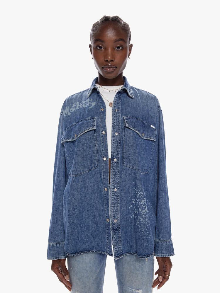 Front view of woman in an oversized denim button up with long sleeves, oversized patch pockets, a curved hem and snaps down the front made from recycled cotton blend in a midblue wash with mothers name beached in death metal inspired front on the chest and back