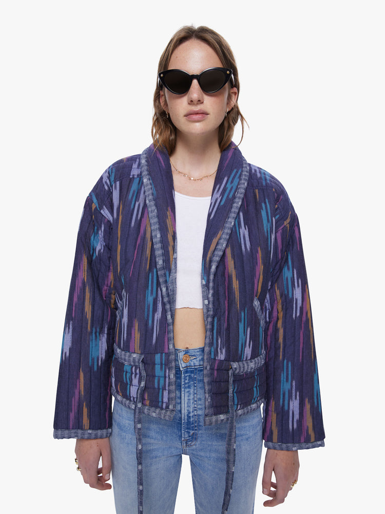 Front view of woman in a shawl collar, drop shoulders, dark blue hue quilted jacket with brush stroke inspired pattern in lavender, mustard, and baby blue that has a drawstring waist