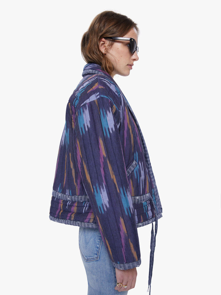 Side view of woman in a shawl collar, drop shoulders, dark blue hue quilted jacket with brush stroke inspired pattern in lavender, mustard, and baby blue that has a drawstring waist