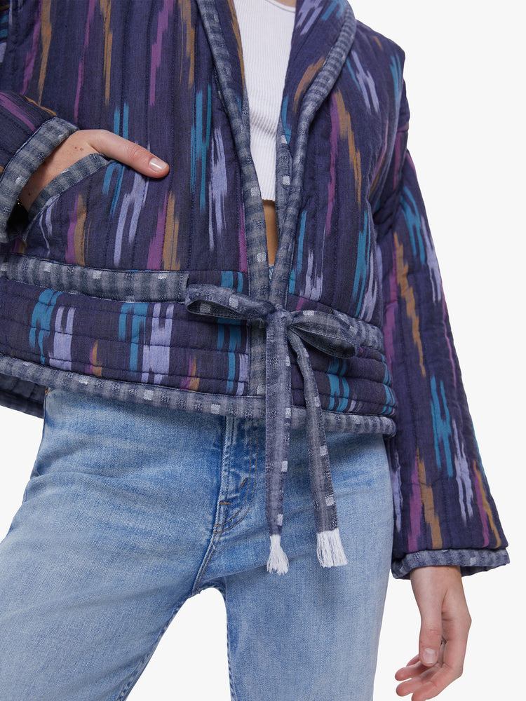 Close up view of woman in a shawl collar, drop shoulders, dark blue hue quilted jacket with brush stroke inspired pattern in lavender, mustard, and baby blue that has a drawstring waist